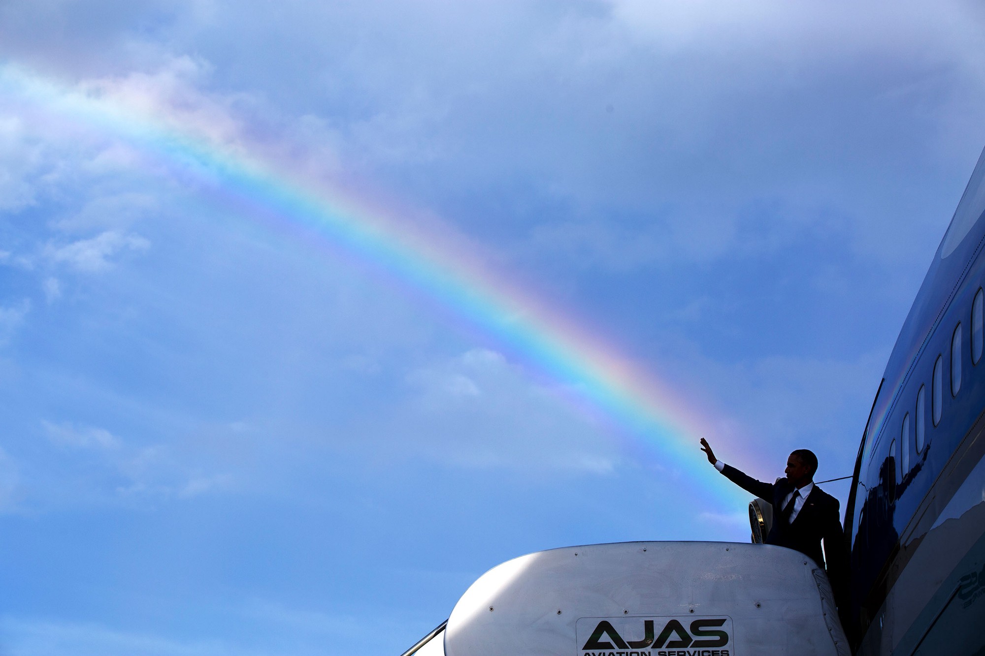 โอบามาโบกมือเป็นจังหวะซ้อนกับสายรุ้งพอดีขณะขึ้นเครื่องแอร์ฟอร์ซวันที่สนามบินนานาชาตินอร์แมน แมนลีย์ ในกรุงคิงสตัน ประเทศจาเมกา เมื่อ 9 เมษายน 2558 / Official White House Photo by Pete Souza