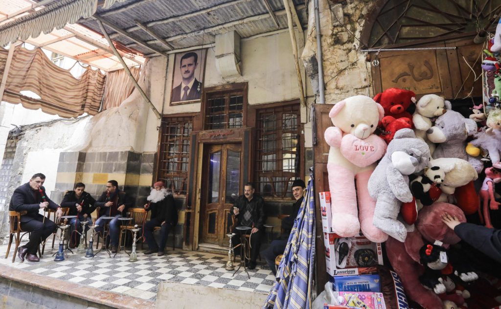 TOPSHOT - Syrians sit outside a cafe bearing the image of President Bashar al-Assad as a shop nearby sells Valentine's Day gifts, in the old part of the capital Damascus on February 12, 2017. / AFP PHOTO / Louai Beshara