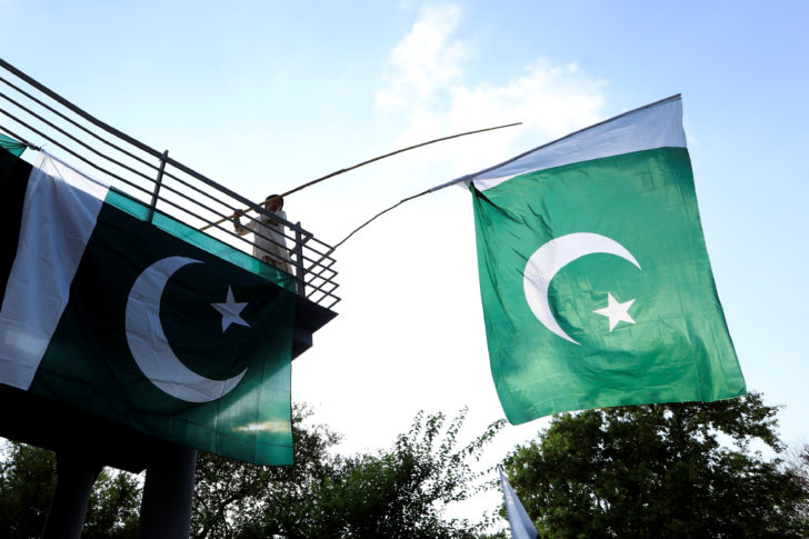 A boy uses a bamboo stick to adjust national flags at an overhead bridge ahead of Pakistan's Independence Day, in Islamabad