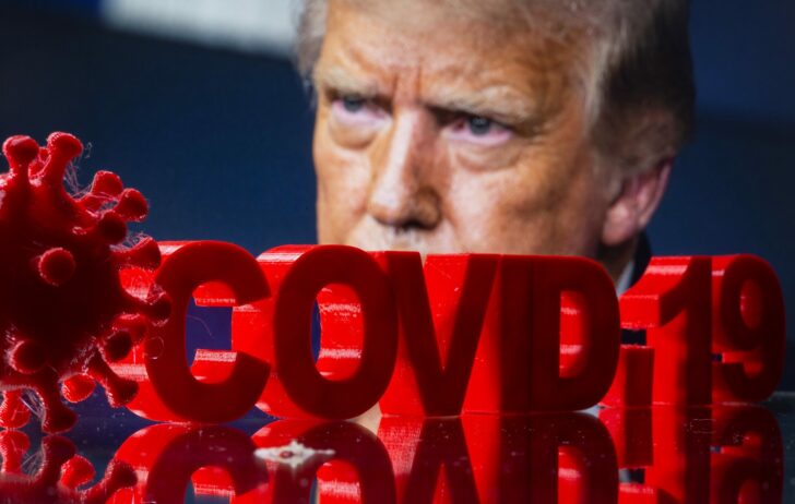 3D printed COVID-19 sign and coronavirus model are seen in front of image of U.S. President Trump