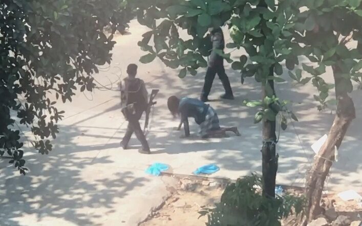 Police officer carrying a rifle forces a man to crawl on all fours in Yangon