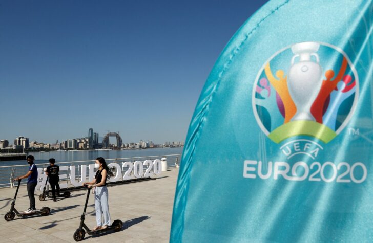 Young people ride scooters past a giant EURO2020 hashtag at a compound of the official fan zone in central Baku