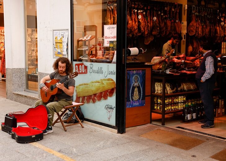 A street musician plays a guitar as a worker cuts ham in a shop in Ronda